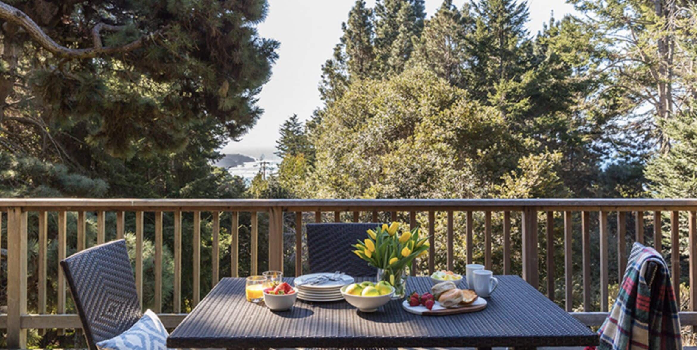 Una terraza de madera con una mesa dispuesta para el desayuno, rodeada de sillas, con vistas a una zona boscosa.