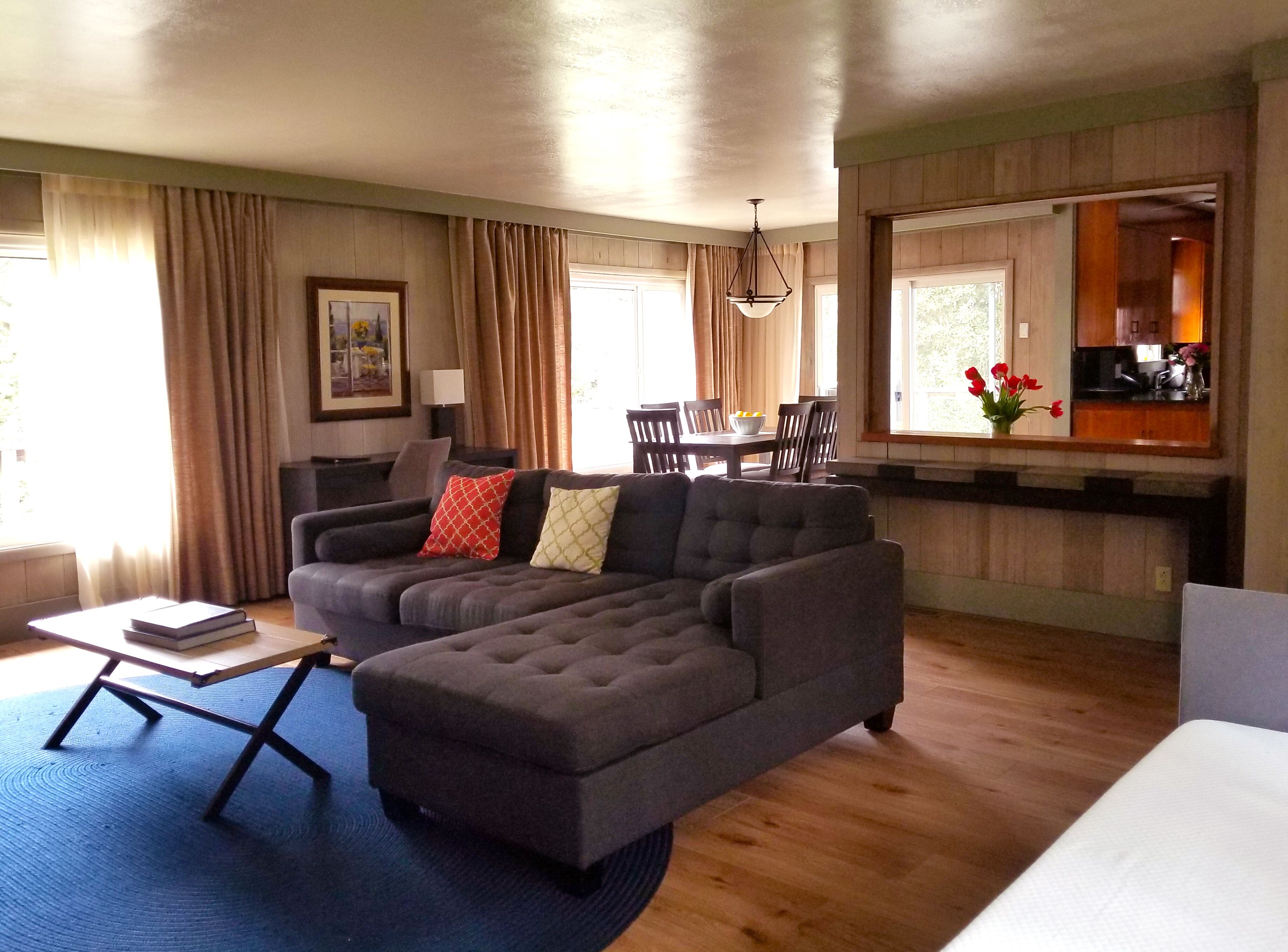 Ein Wohnzimmer mit Ecksofa, Esstisch mit Stühlen, Küchenbereich und großem Fenster mit Vorhängen.