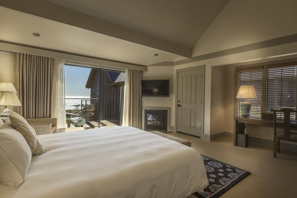 A hotel room with a large bed, a fireplace, a balcony with a view, and a dining area with a table and chairs.