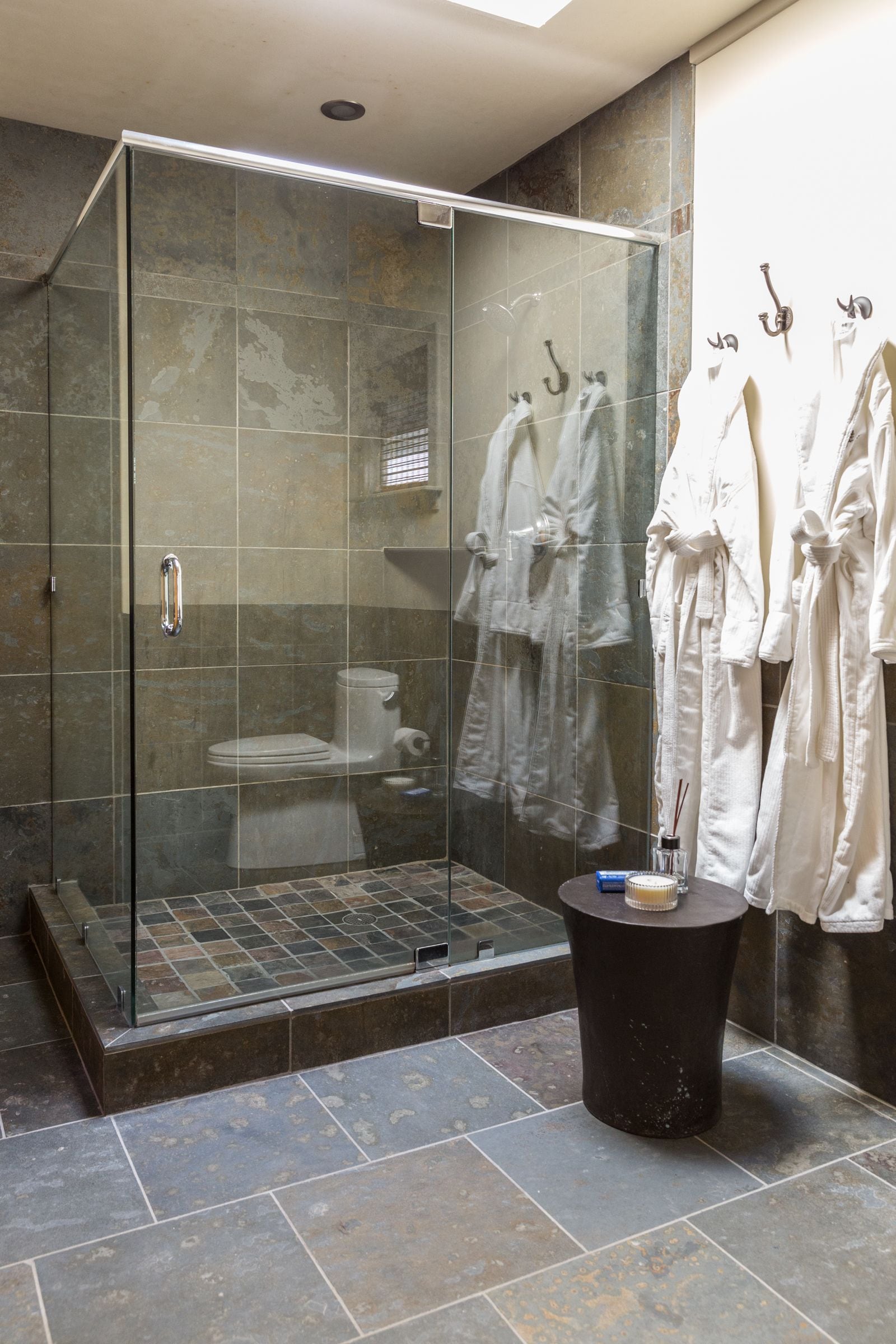 A glass-enclosed shower with a tiled floor, a black pedestal table with a bottle and a brush, and white robes hanging on hooks.