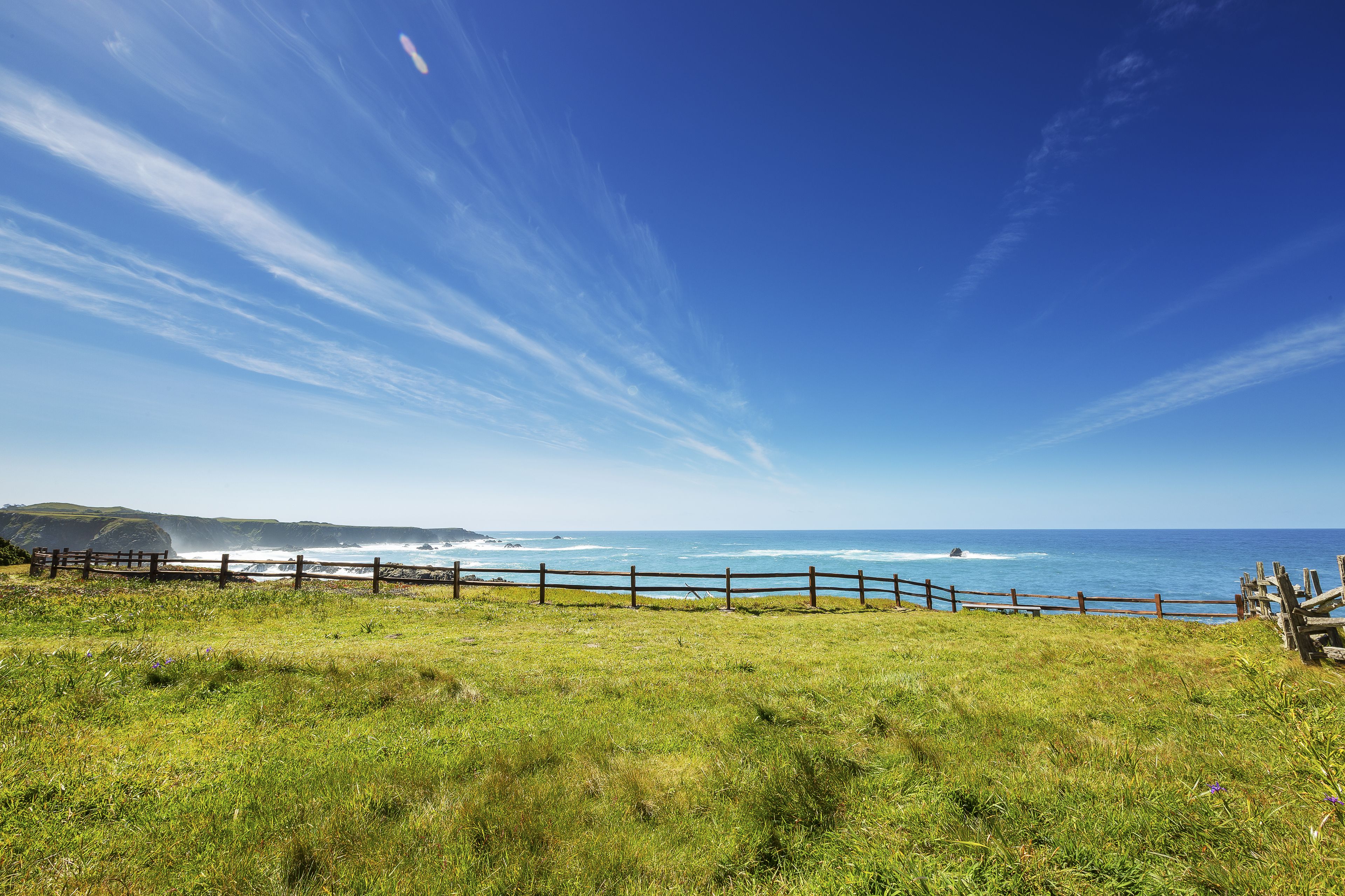 A coastal landscape with a wooden fence, grassy field, and ocean view.