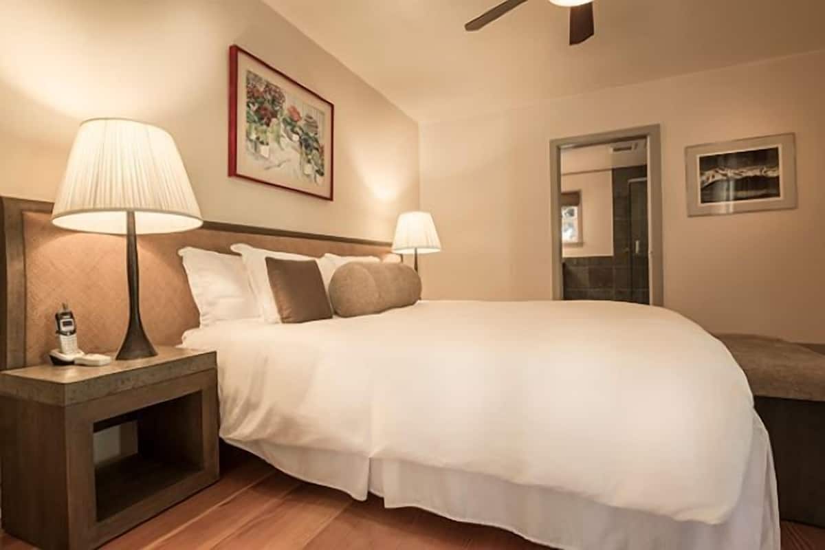 A hotel room with a bed, two bedside lamps, a nightstand, a framed picture on the wall, and a door leading to another room.