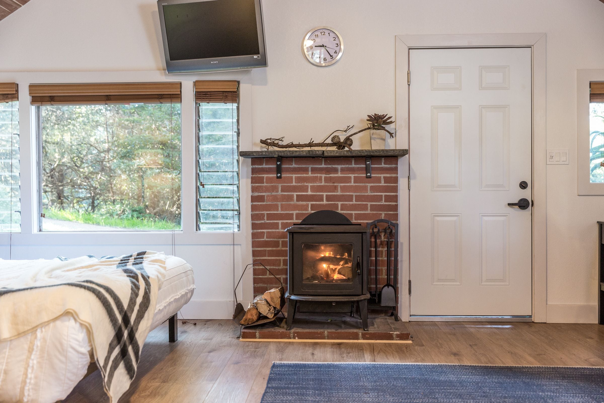 A cozy room with a brick fireplace, a bed with a plaid blanket, a mounted TV, and a door with a window view.