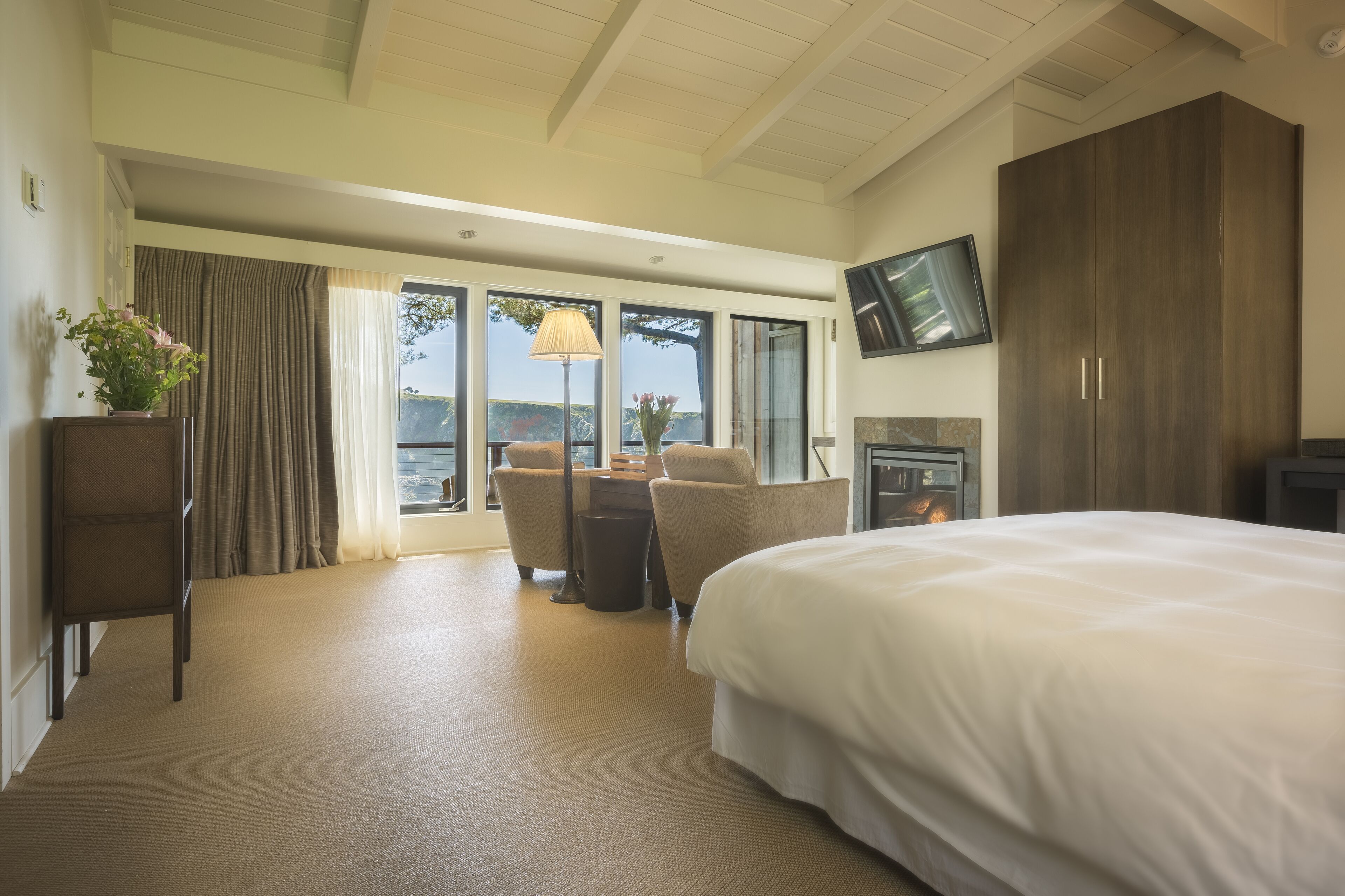 A hotel room with a large bed, a desk with a lamp, a chair, a TV mounted on the wall, and a view of the outdoors through the windows.