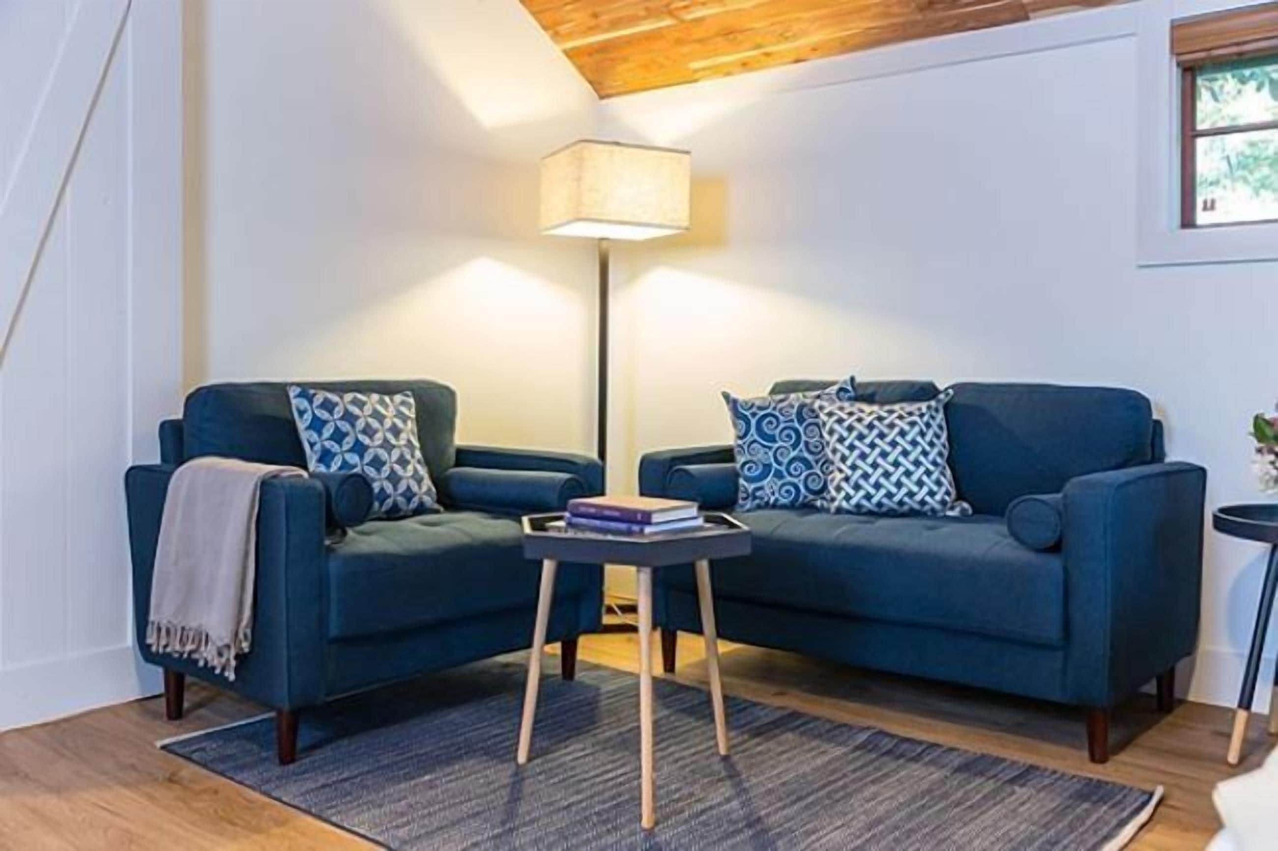 A cozy living room with two blue sofas, patterned cushions, a small side table with books, a floor lamp, and a window with a view.