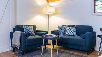 A cozy living room with two blue sofas, patterned cushions, a small side table with books, a floor lamp, and a window with a view.