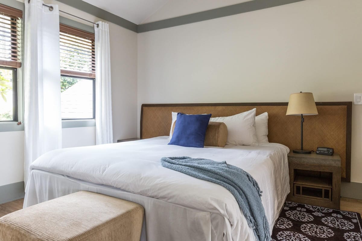 A neatly made bed with white bedding and a blue pillow, a bedside table with a lamp, and a window with curtains.