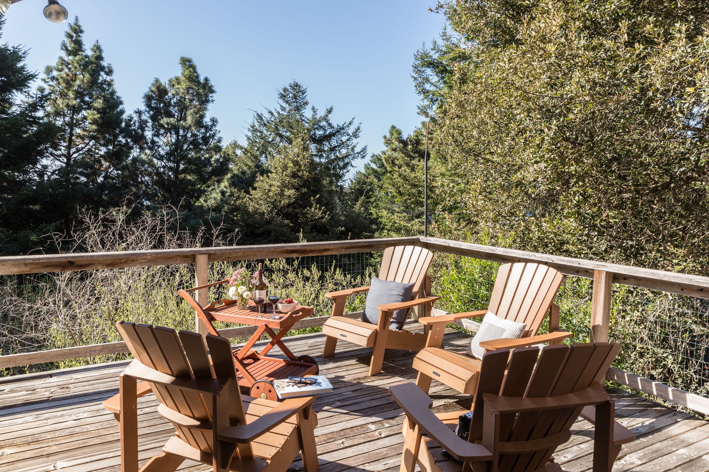A wooden deck with Adirondack chairs and a table, surrounded by trees and a clear sky.