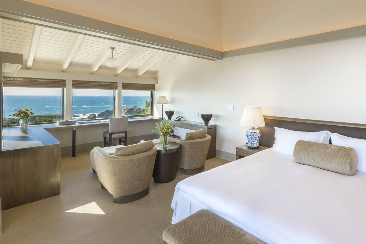 A hotel room with a large bed, two armchairs, a desk, and a view of the ocean.