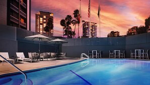 A swimming pool with lounge chairs and umbrellas, surrounded by buildings and palm trees under a sunset sky. - The DEXTRO Little Italy, BW Premier Collection (San Diego)