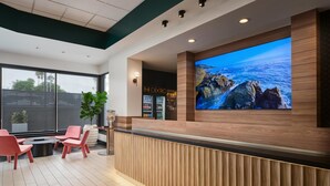 A modern reception area with a wooden reception desk, a large flat-screen TV displaying a scenic ocean view, and a seating area with pink chairs. - The DEXTRO Little Italy, BW Premier Collection (San Diego)