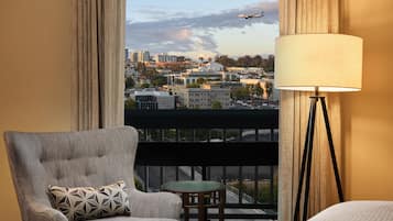 A hotel room with a view of a cityscape, a chair with a patterned pillow, a small round table, and a floor lamp.