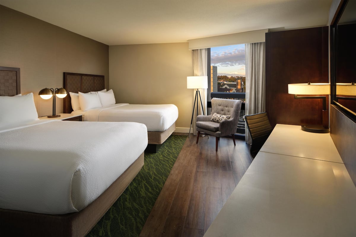 A hotel room with two beds, a desk, a chair, a lamp, and a view of the city.