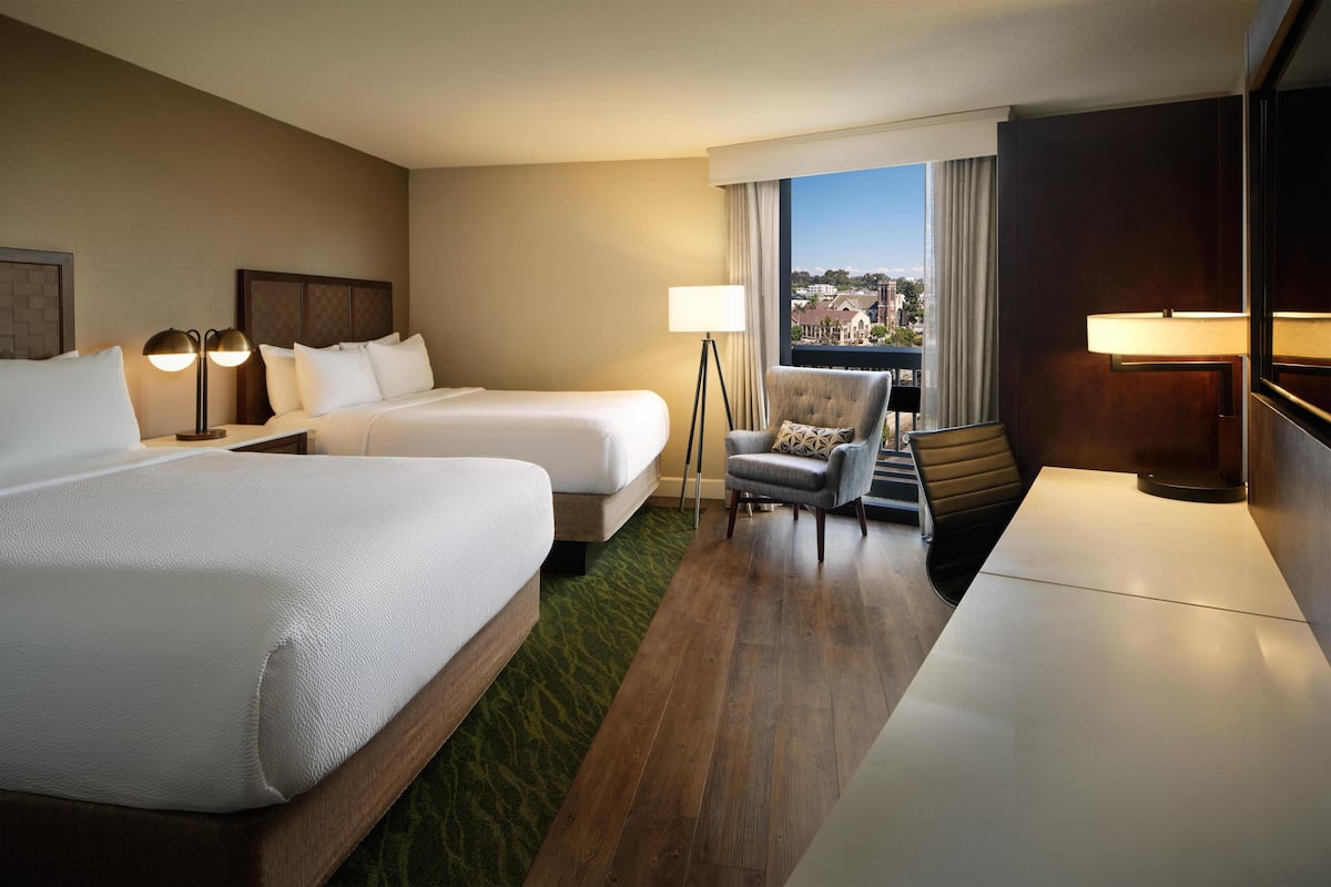 A hotel room with two beds, a chair, a desk, and a view of a cityscape.