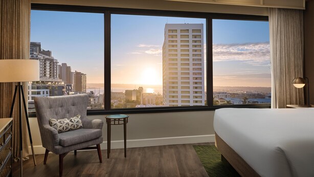 A hotel room with a large window offering a city view, a bed, a chair, and a small table. - The DEXTRO Little Italy, BW Premier Collection (San Diego)