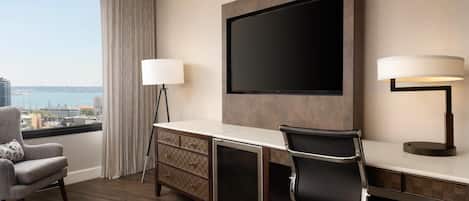 A hotel room with a large window, a desk with a flat-screen TV, two lamps, a chair, and a dresser.