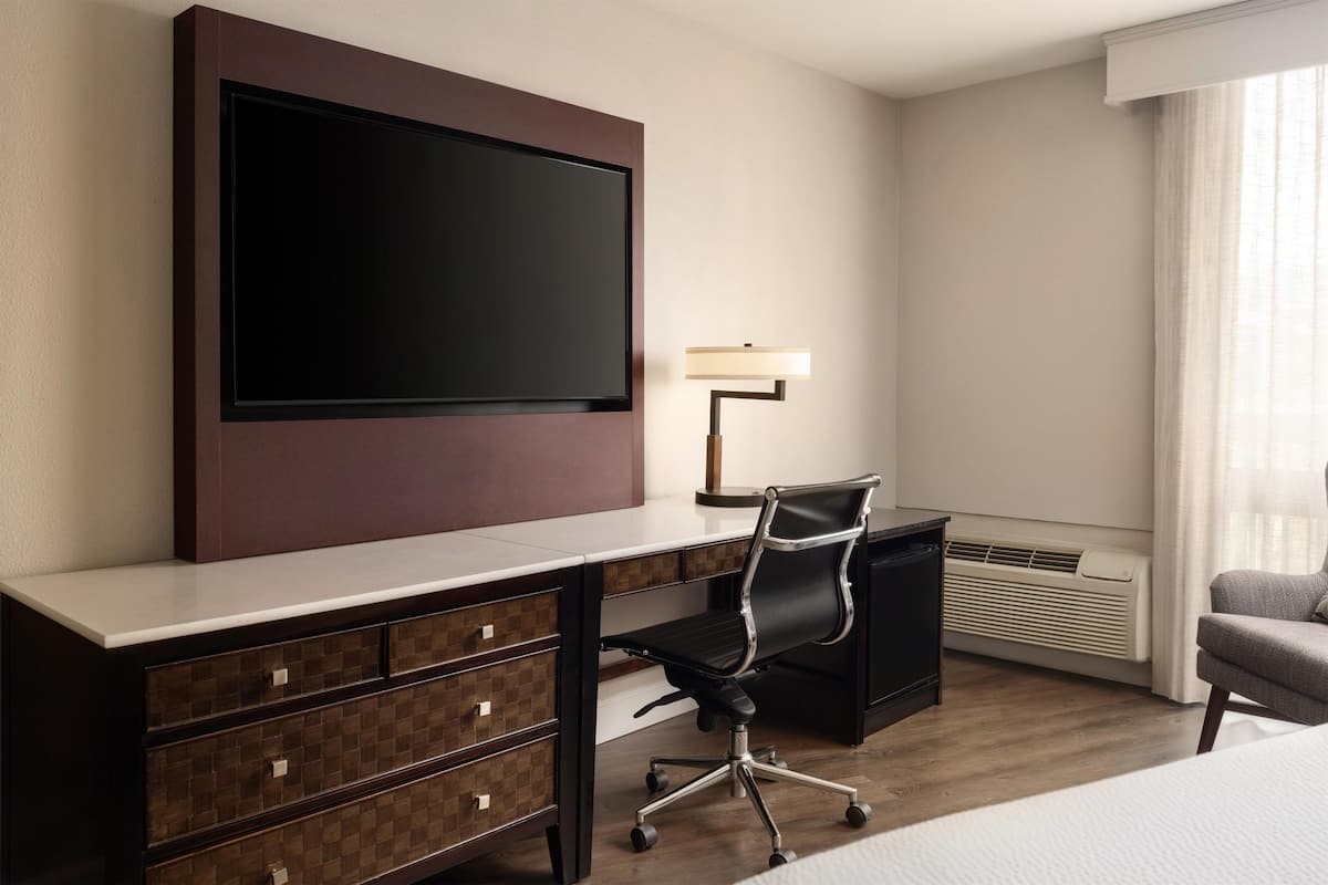 A hotel room with a large flat-screen TV mounted on the wall, a desk with a chair, a lamp, and a bed.