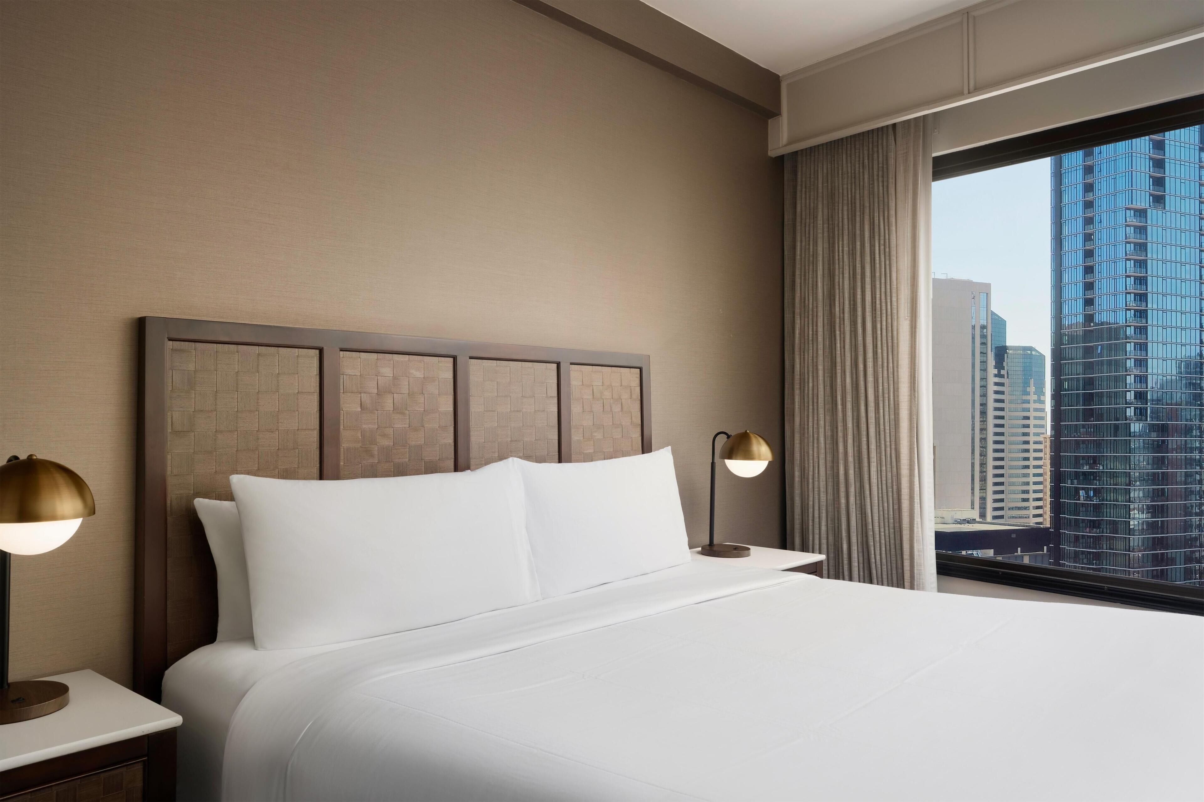 A hotel room with a large bed, two bedside lamps, a view of tall buildings through a window, and a textured headboard.