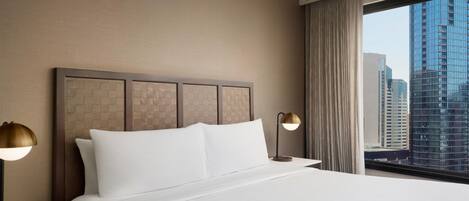 A hotel room with a large bed, two bedside lamps, a view of tall buildings through a window, and a textured headboard.