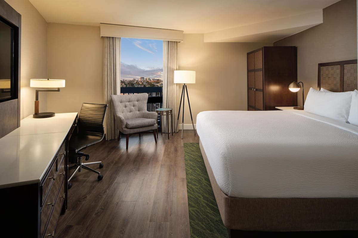 A hotel room with a bed, desk, chair, lamp, and a view of the city through the window.