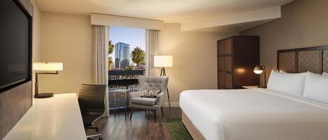 Habitación de hotel con una cama grande, un escritorio, una silla, televisión y vistas a palmeras y edificios.