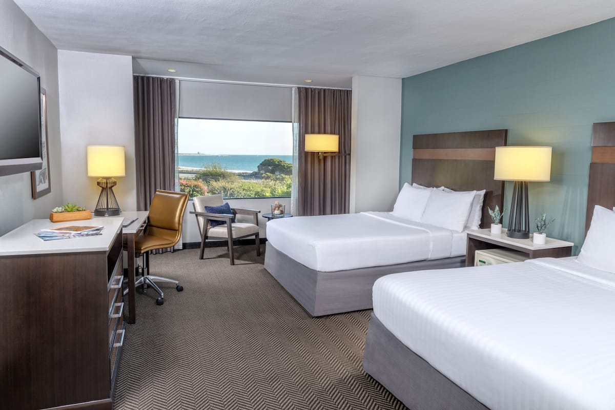 A hotel room with two beds, a desk, a chair, and a large window offering a view of the sea.