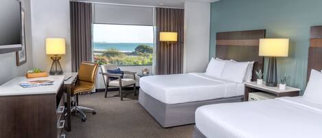 Habitación de hotel con dos camas, un escritorio, una silla y un ventanal con vistas al mar.