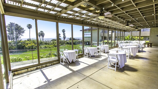 A covered outdoor dining area with tables and chairs, large windows offering a view of greenery and a building in the background. - The Pierpont Inn (Ventura)
