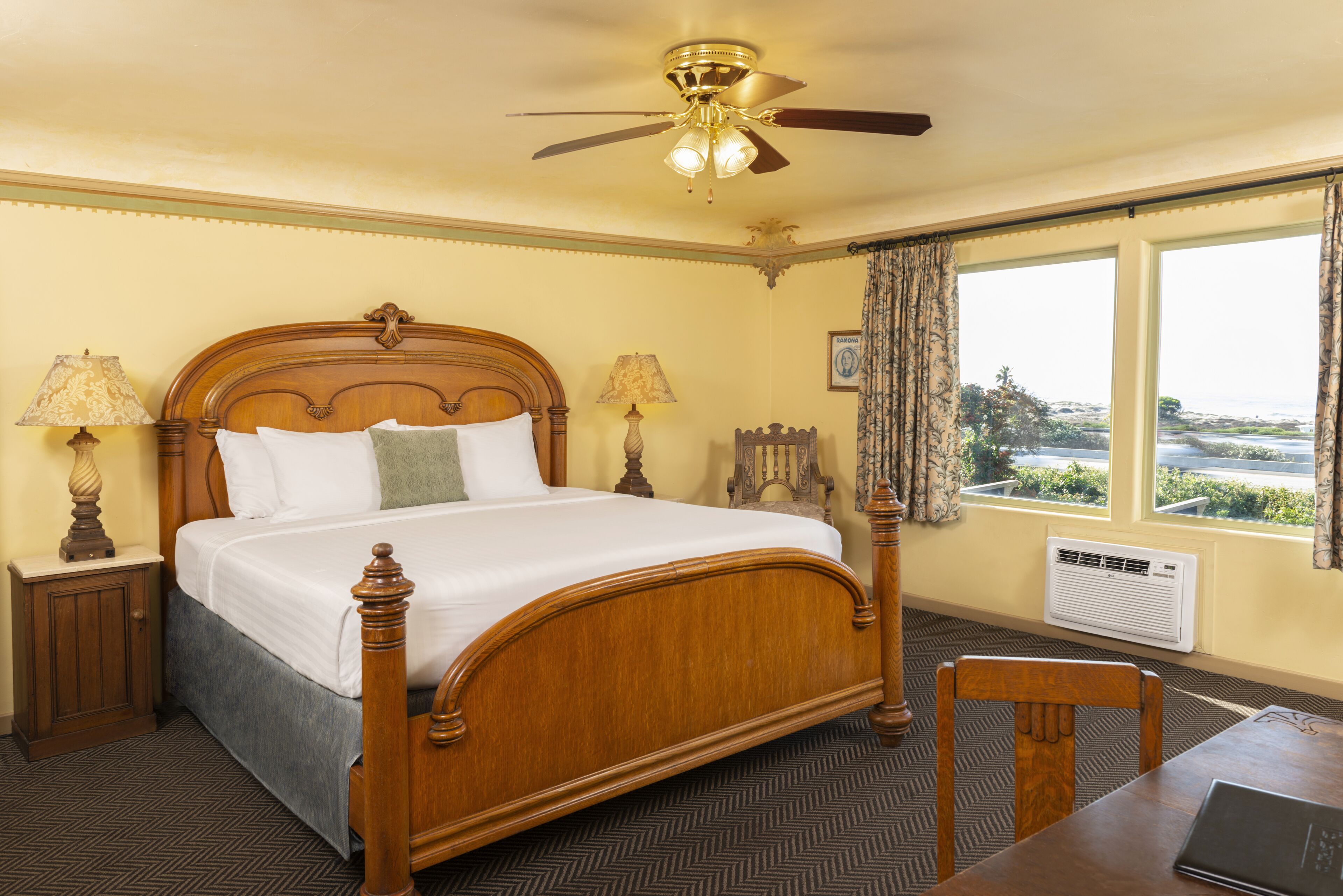A hotel room with a wooden four-poster bed, two bedside lamps, a ceiling fan, a large window with curtains, and a table with a laptop.