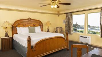 A hotel room with a wooden four-poster bed, two bedside lamps, a ceiling fan, a large window with curtains, and a table with a laptop.