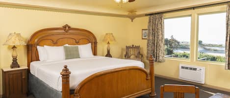 Habitación de hotel con una cama con dosel de madera, dos lámparas de noche, un ventilador de techo, un ventanal con cortinas y una mesa con una computadora portátil.