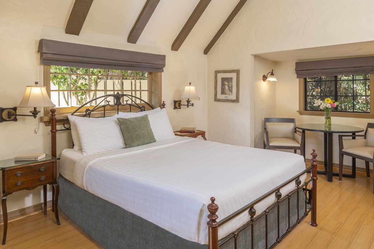 A neatly made bed with a wooden headboard, a bedside table with a lamp, and a chair at a small round table with a vase of flowers.