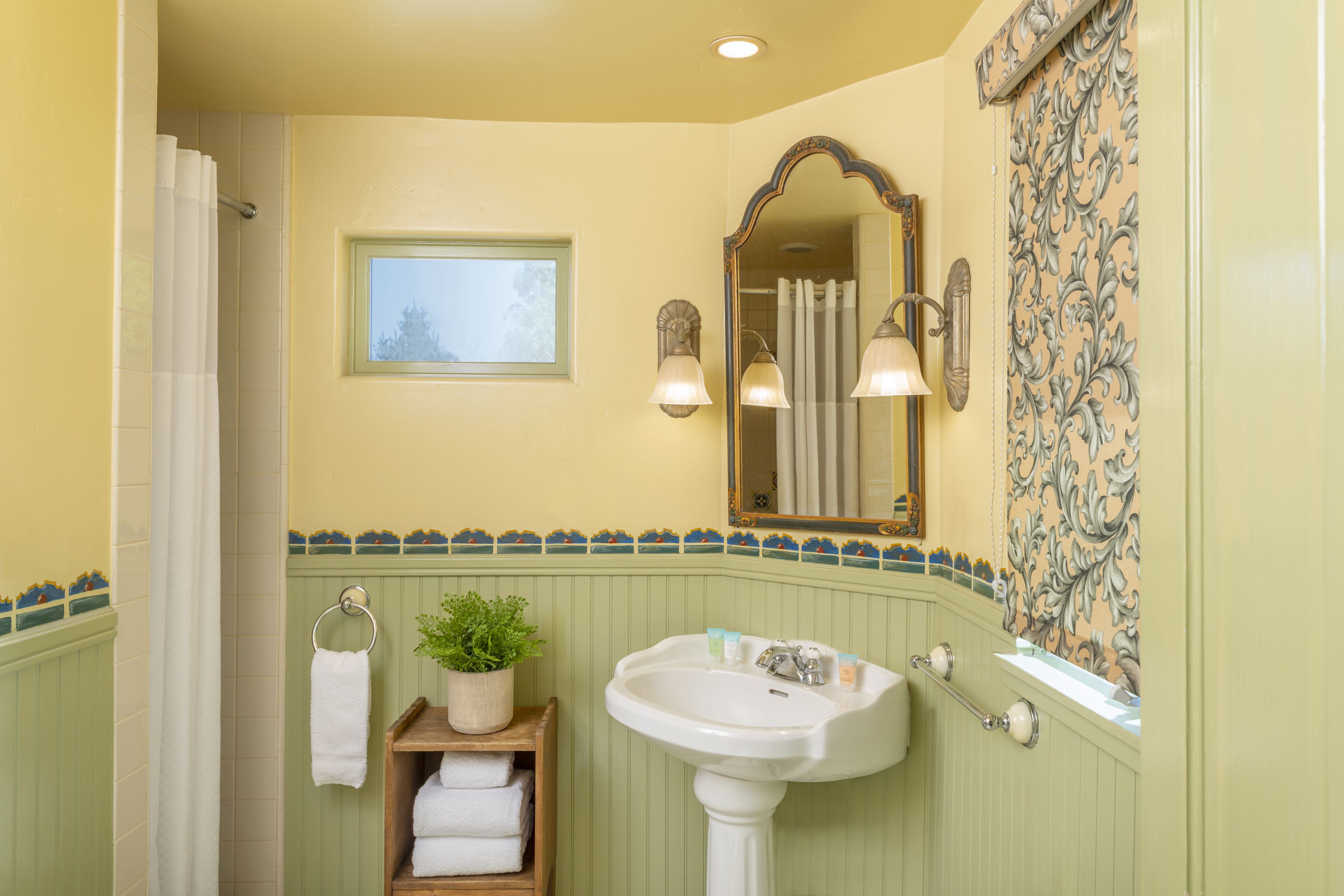 Un baño con lavamanos pedestal, espejo, ventana y perchero para toallas.