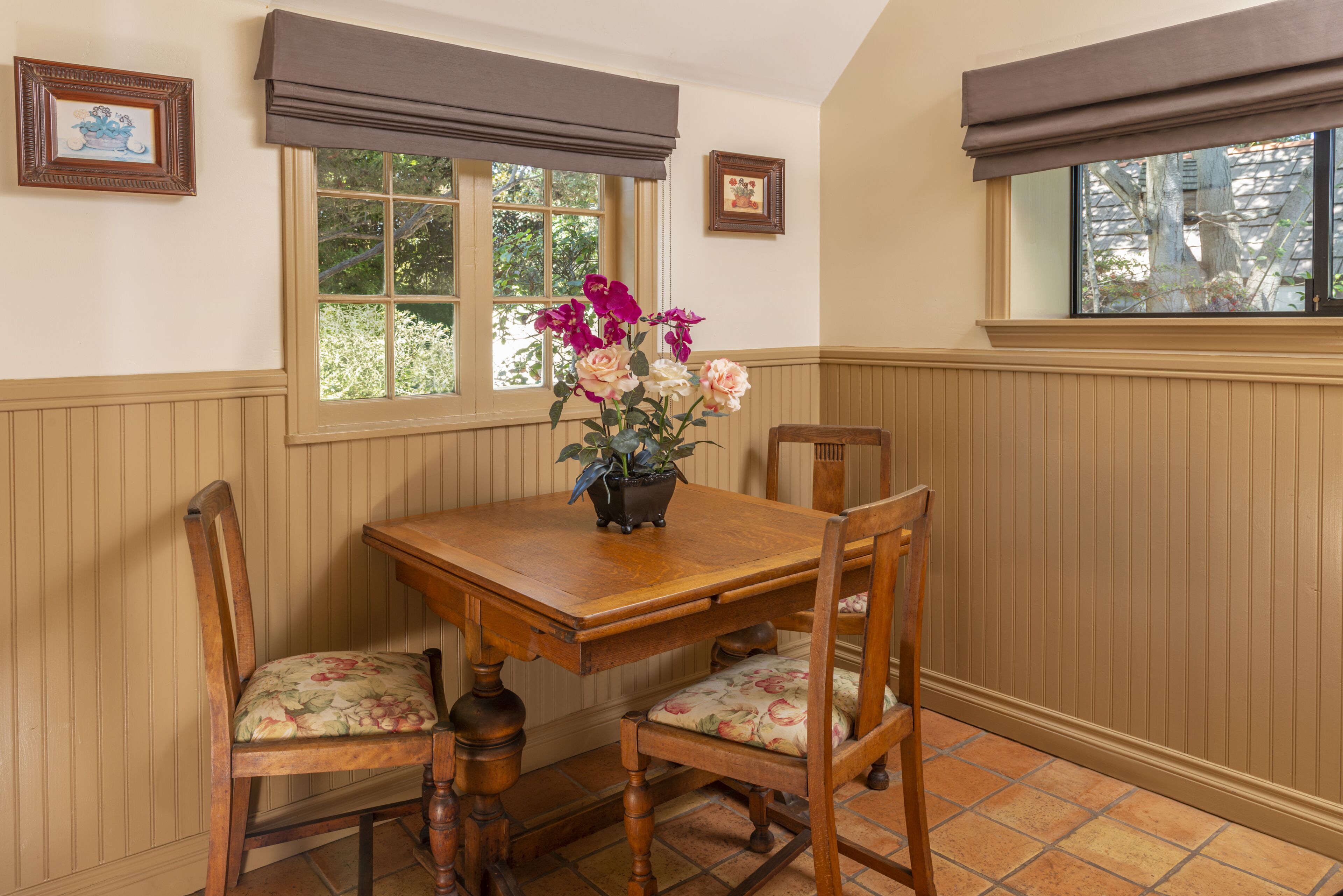 A dining area with a wooden table, floral chairs, a vase of flowers, and a window with blinds.
