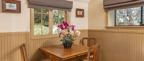 A dining area with a wooden table, floral chairs, a vase of flowers, and a window with blinds.