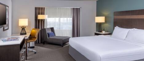 A hotel room with a large bed, a desk with a chair, two lamps, a TV, and a window with curtains.