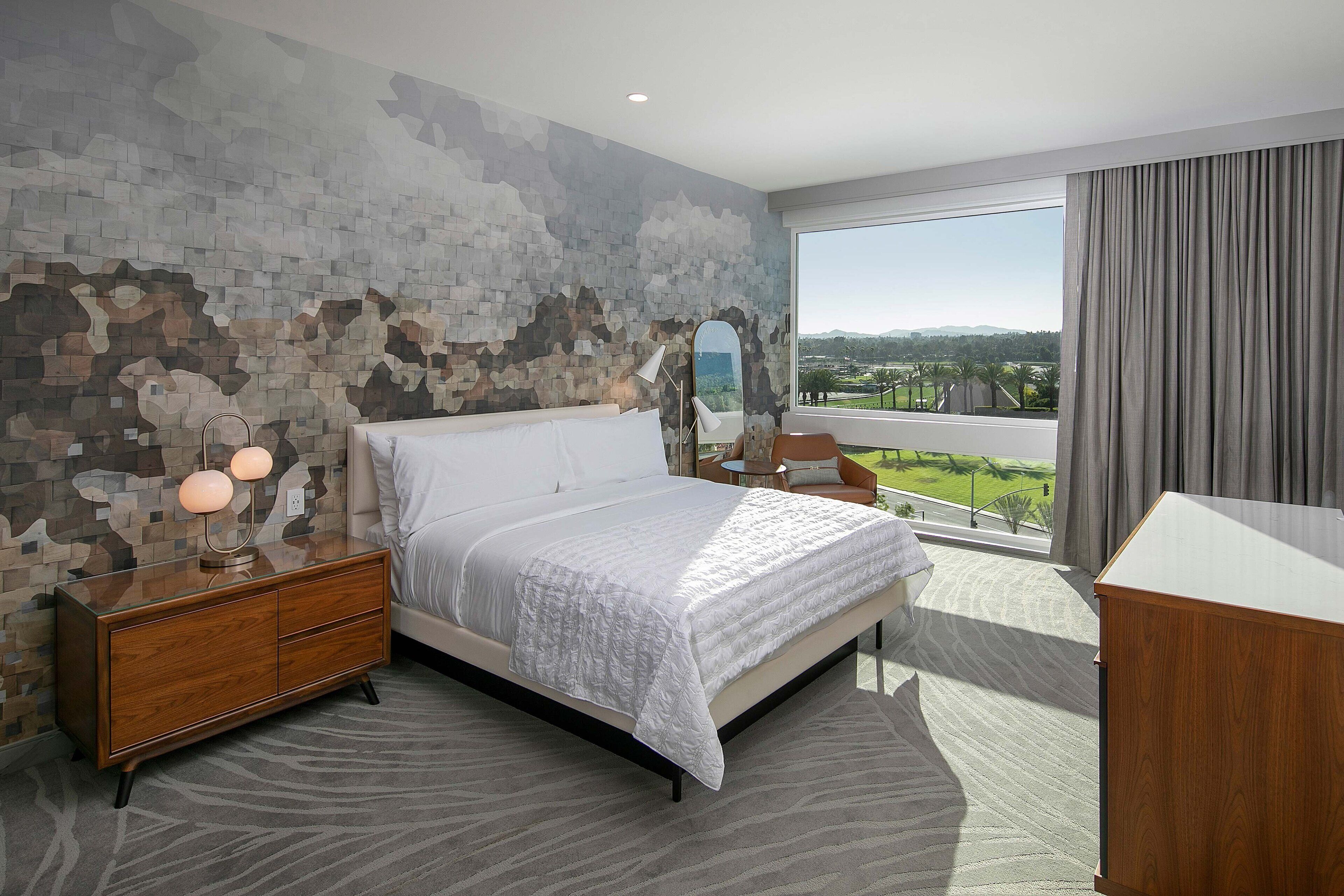 A modern bedroom with a large bed, a wooden dresser, a mirror, and a view of the outdoors.