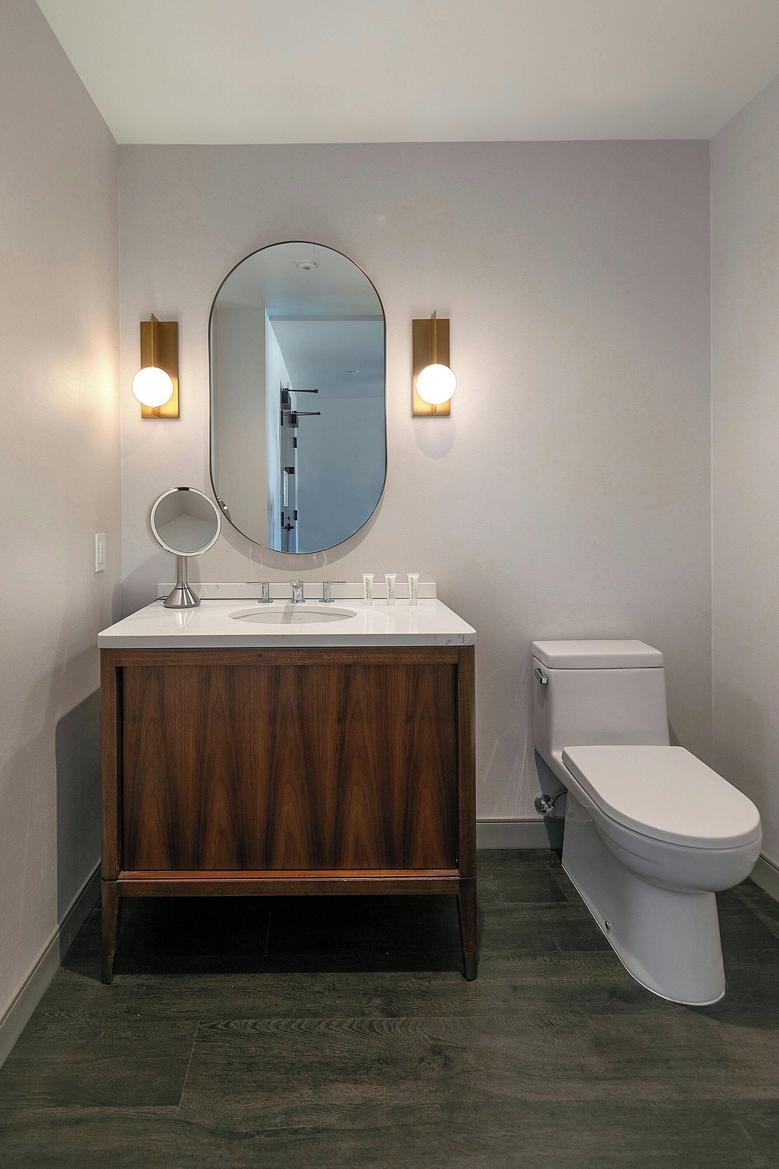 A modern bathroom with a wooden vanity, two wall-mounted lights, a round mirror, and a white toilet.