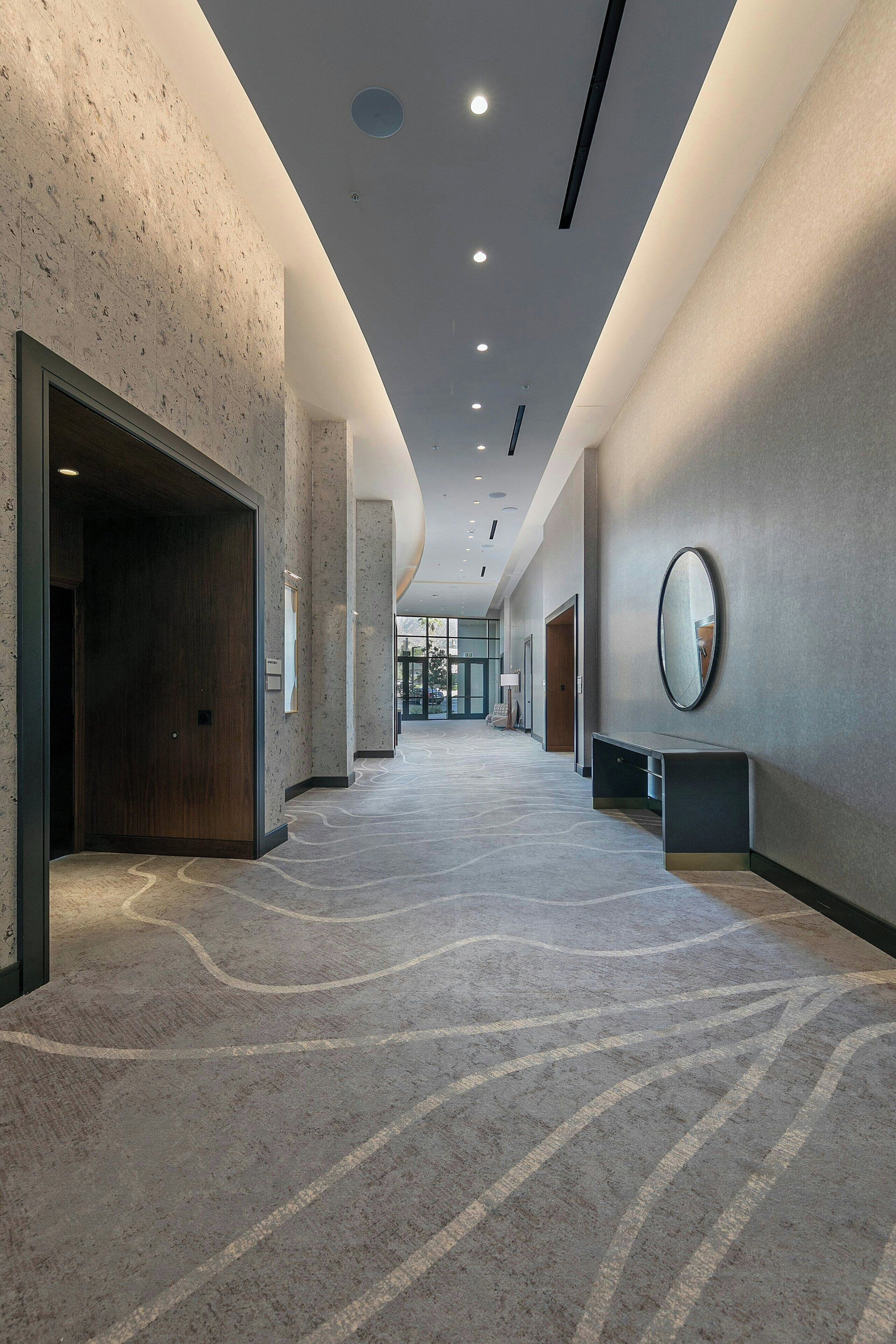 A modern hotel corridor with a patterned carpet, a wooden door, a bench, and a mirror on the wall.