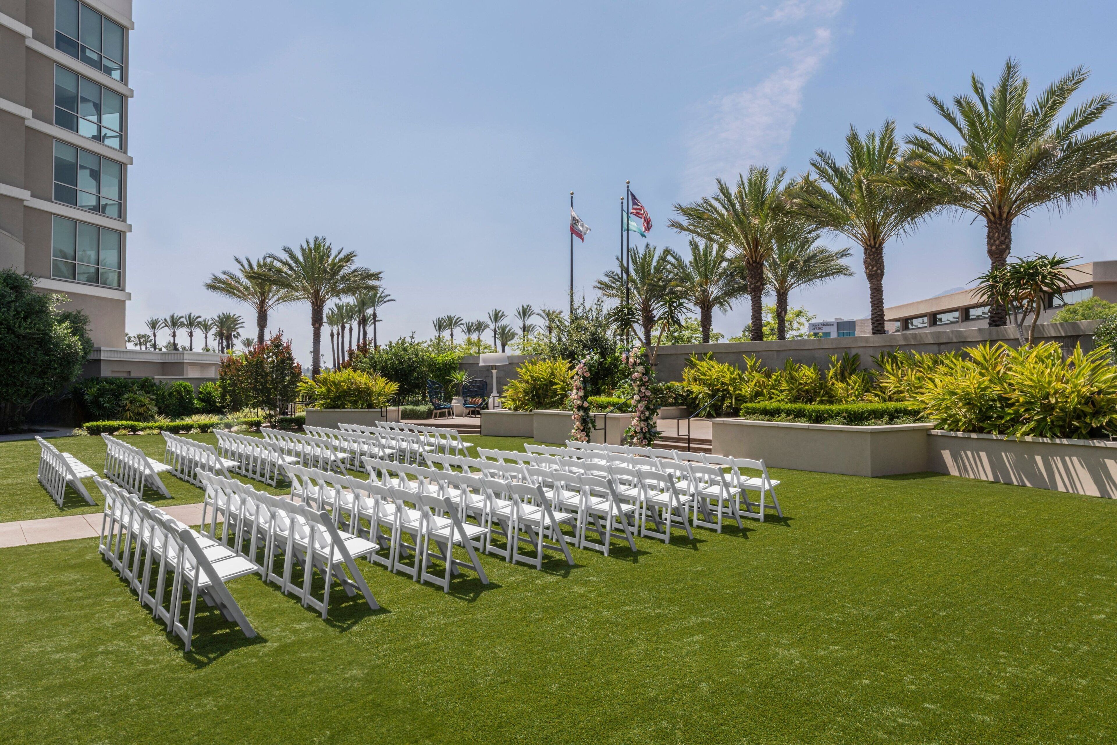 A well-maintained outdoor area with rows of white chairs arranged for an event, surrounded by lush greenery and palm trees.