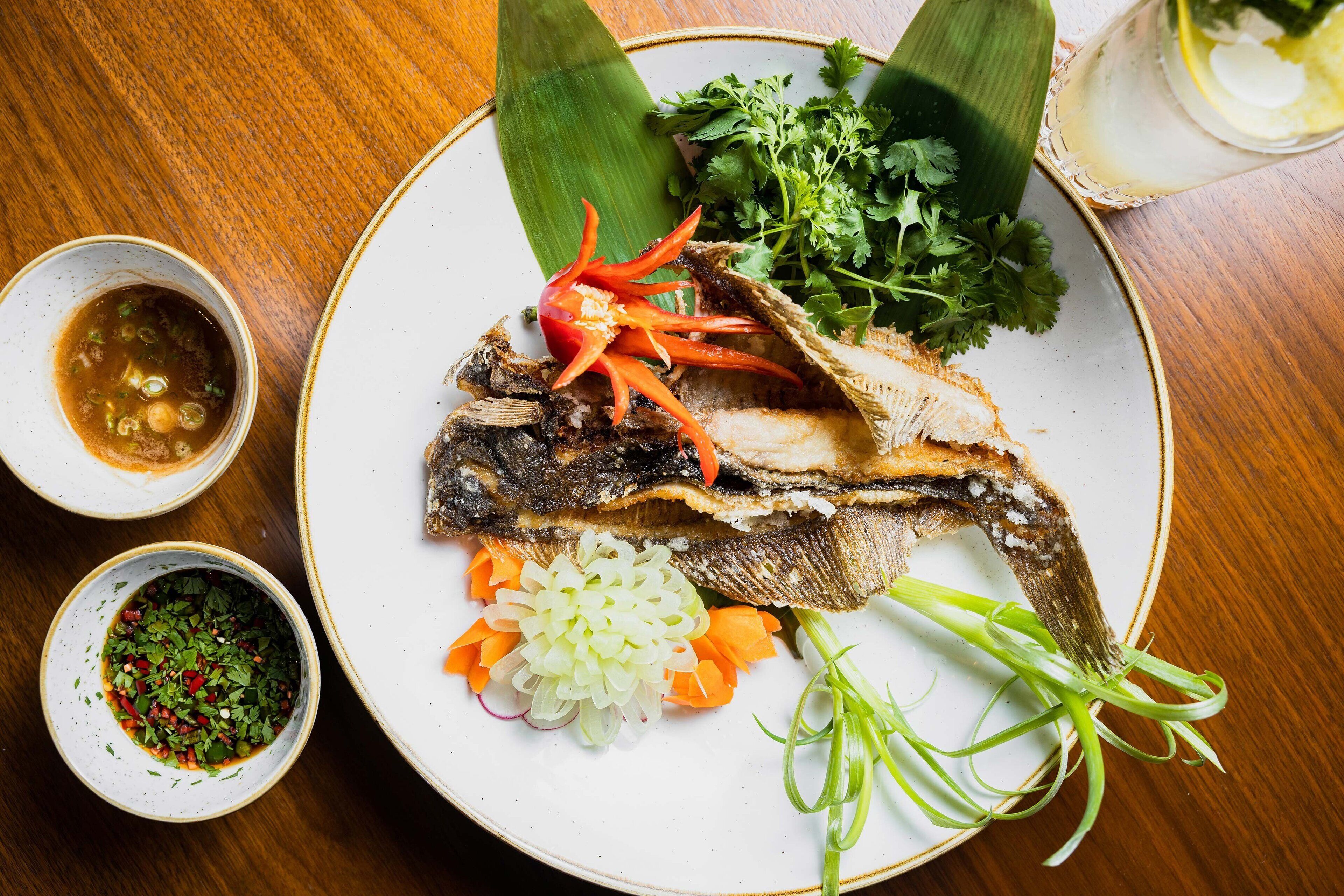 A plate with a whole fish, garnished with herbs and vegetables, and two small bowls of dipping sauces.