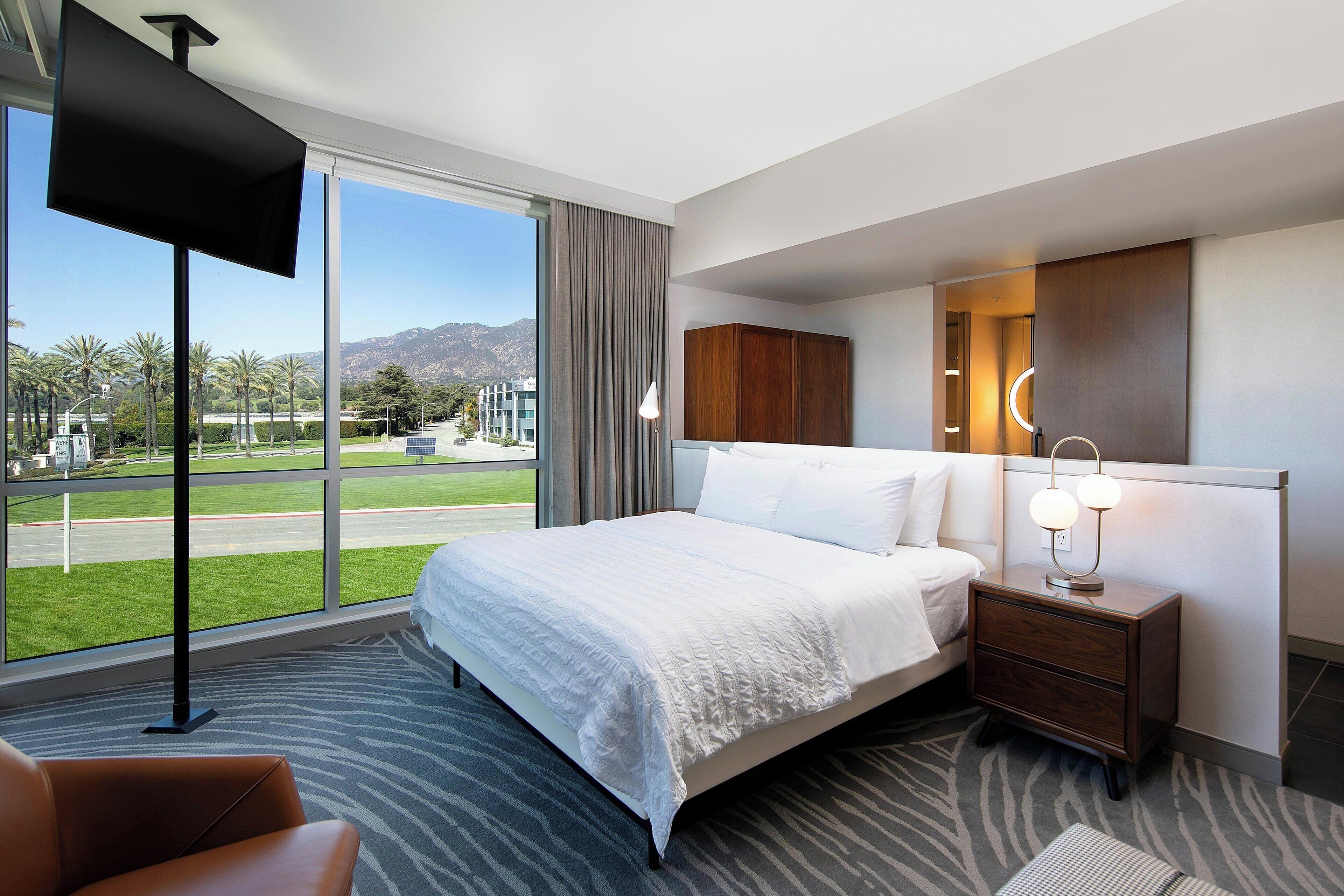 A modern hotel room with a large bed, a nightstand with a lamp, a view of the city and mountains, and a large window.