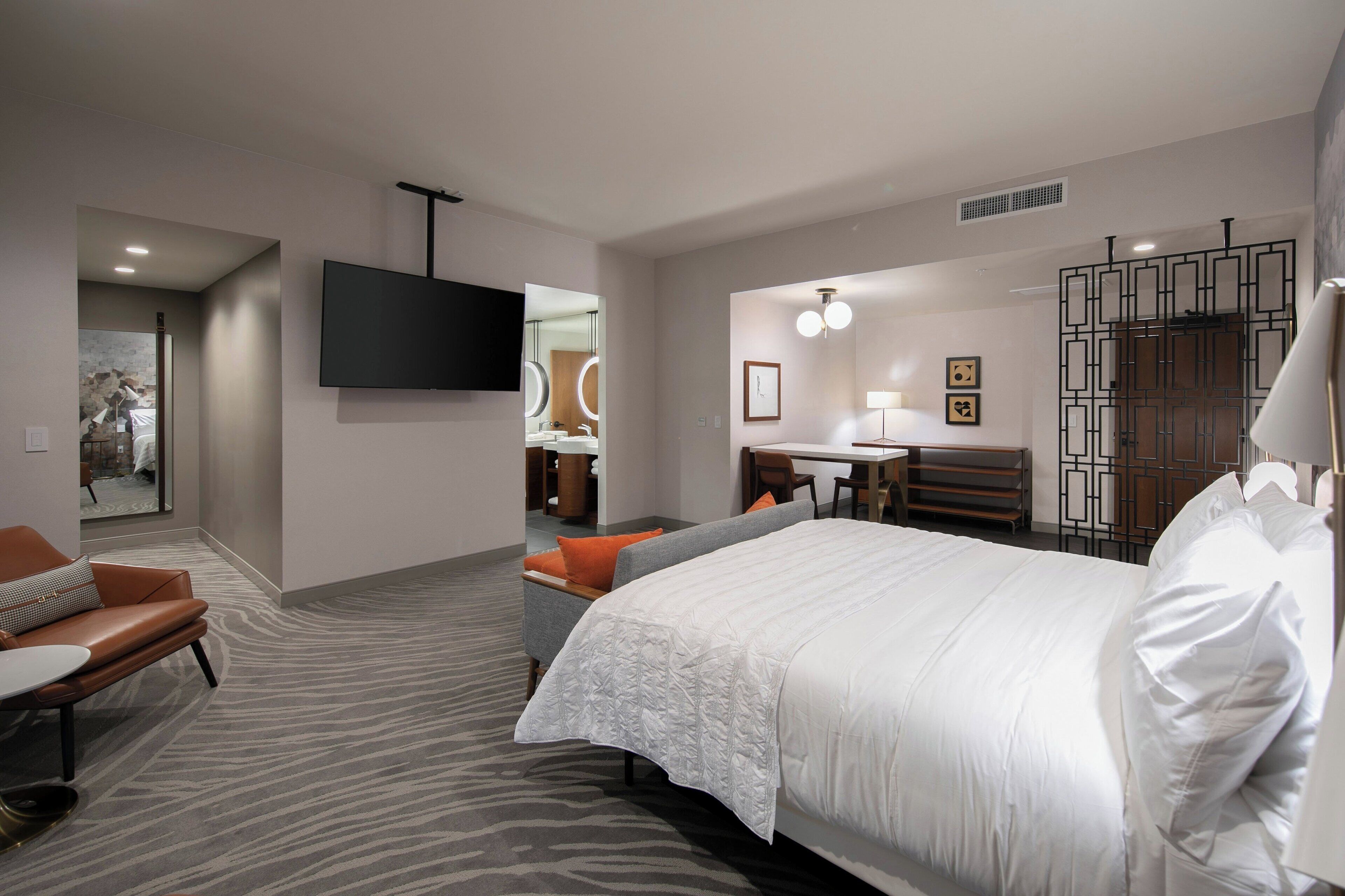 Ein modernes Hotelzimmer mit einem großen Bett, einer Sitzecke mit Sofa und Sesseln, einem Esstisch und einem an der Wand befestigten Fernseher.