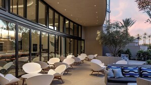 A modern outdoor seating area with white chairs and a sofa, large windows, and a view of palm trees. - Le Méridien Pasadena Arcadia (Arcadia)