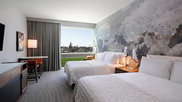 A hotel room with two beds, a desk, and a chair. There is a large window with curtains, a wall with a mosaic design, and a view of the outdoors.