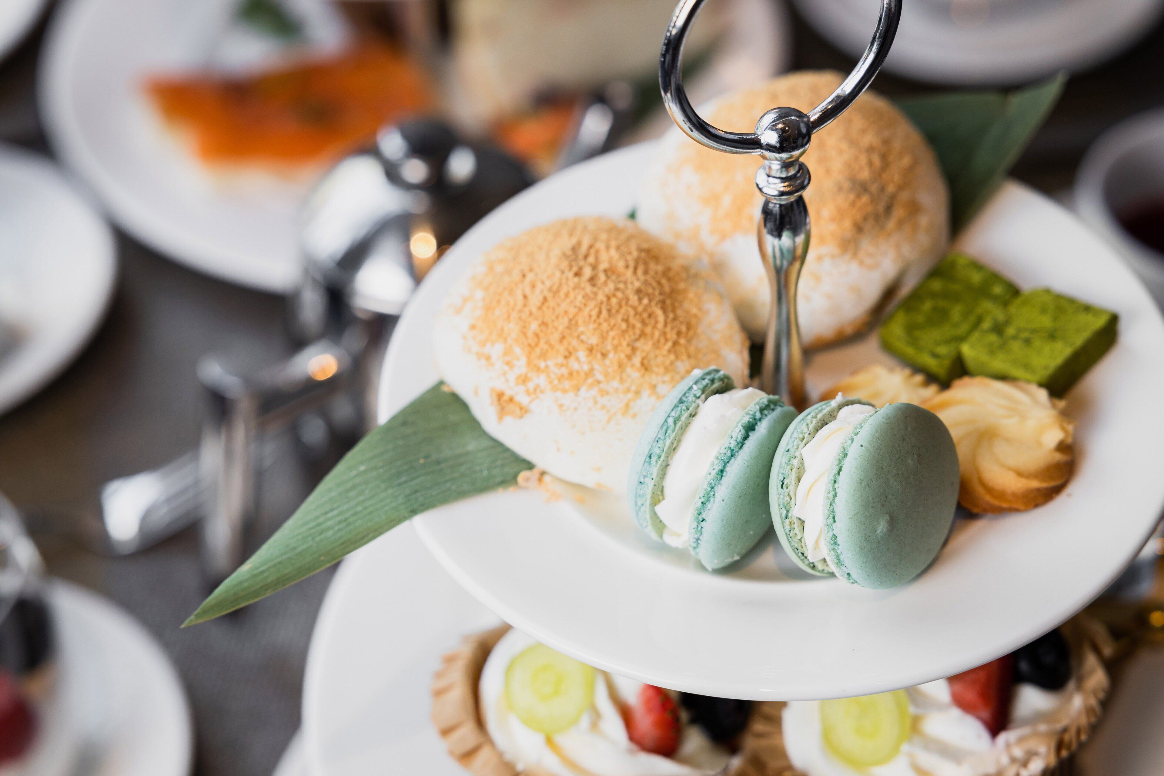A tiered dessert plate with macarons, a cake, and a green dessert, set on a table with other dishes and utensils.