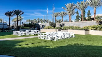 Un espacio al aire libre cubierto de césped, con filas de sillas blancas dispuestas para un evento, palmeras y un asta con banderas.