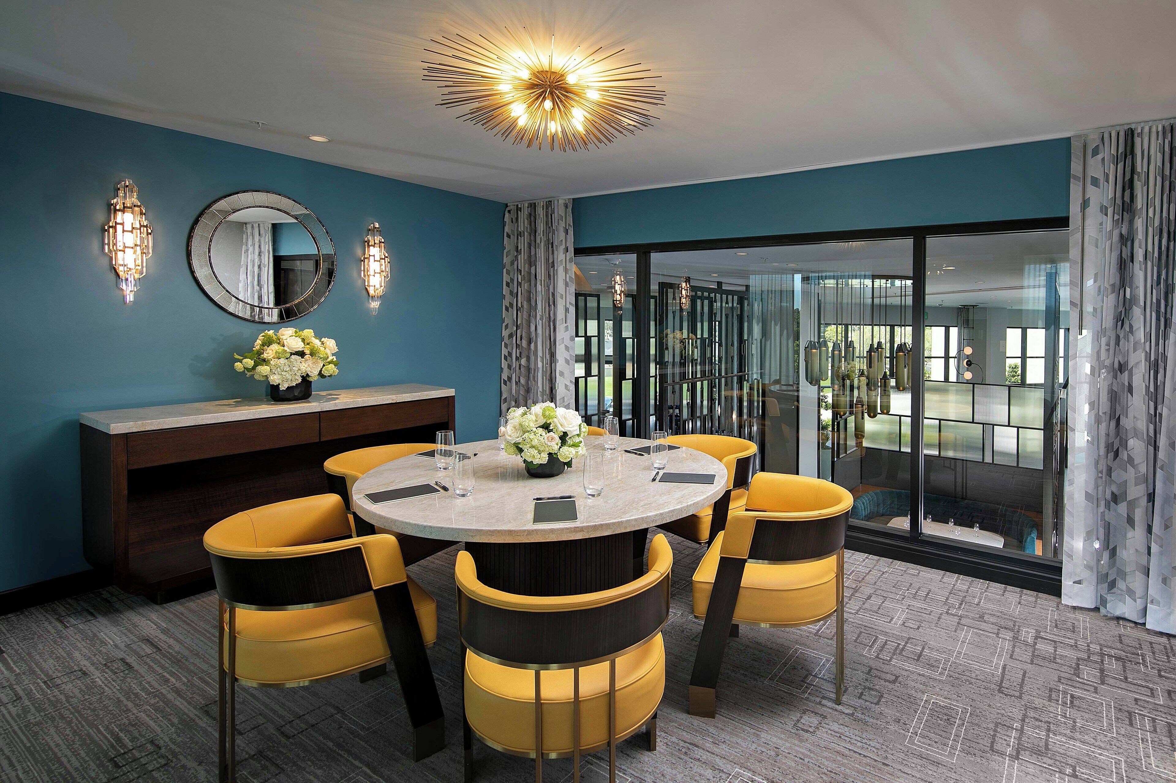 Uma sala de jantar moderna com uma mesa redonda, cadeiras amarelas, um vaso de flores e um espelho grande na parede.