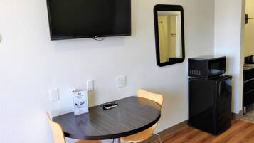 Een kleine kamer met een donker houten tafeltje, twee gele stoelen, een flatscreen-tv aan de muur, een zwarte magnetron en een koelkast.