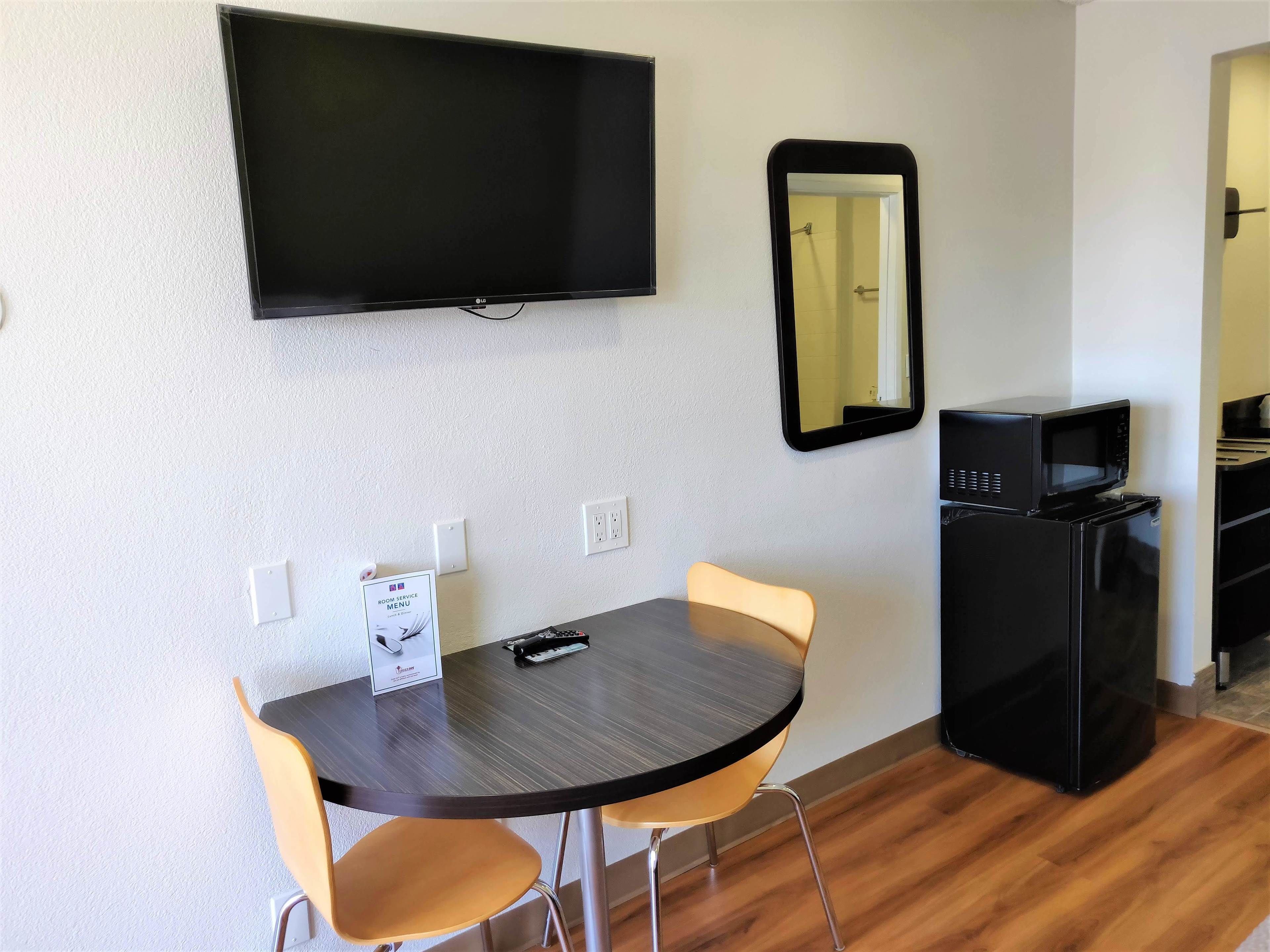 Una habitación pequeña con una mesa de madera oscura, dos sillas amarillas, un televisor negro montado en la pared, un microondas negro y un refrigerador.